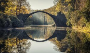 Zu sehen ist die Rakotzbruecke samt Spiegelung im Wasser. Um die Bruecke herum sind Bäume mit gelben Blättern zu sehen.